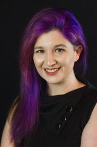 Shoulder-and-up headshot of a white woman with purple hair and mischievous smile. She is wearing a sleeveless black dress.